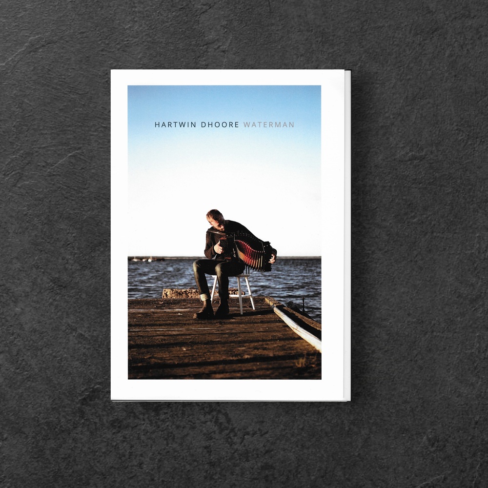 Ein Mann sitzt auf einem weißen Stuhl auf einem Holzsteg am Wasser und spielt ein Akkordeon. Er trägt eine dunkle Jacke, Jeans und Stiefel. Der Himmel ist klar und blau, während das Wasser im Hintergrund ruhig wirkt. Der Name "Hartwin Dhoore" und der Albumtitel "Waterman" sind oben auf dem Bild zu sehen.